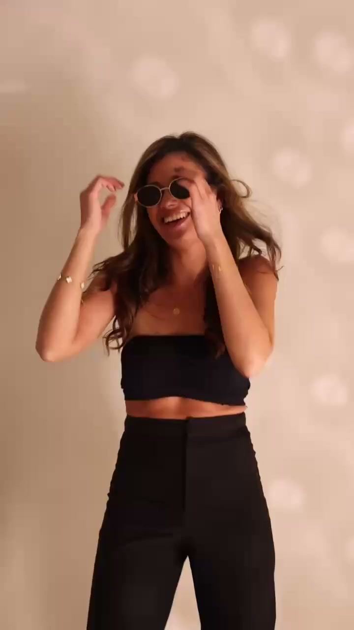 Naomi met zonnebril, lachend en dansend voor een warme, lichte wand.